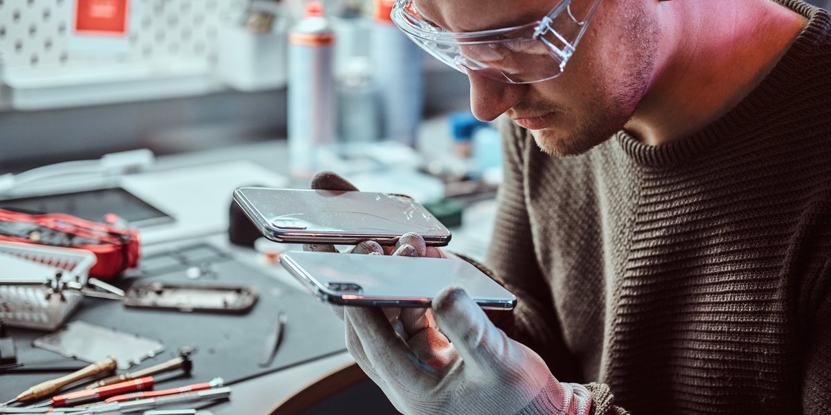 Ein Techniker mit Schutzbrille und weißen Handschuhen repariert ein Smartphone in einer Werkstatt. Er trägt einen braunen Strickpullover und hält das Gerät vorsichtig in beiden Händen, während er es genau untersucht. Auf dem Arbeitstisch sind verschiedene Reparaturwerkzeuge, Schraubendreher und technische Ausrüstung zu sehen. Die Szene zeigt eine professionelle Handy-Reparaturwerkstatt mit konzentrierter Facharbeit.
